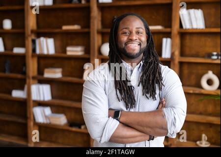 Cheerful African-American modern businessman, a guy with dreadlocks hairstyle stands in contemporary office space or library in confident pose with folded arms, looks at the camera and toothy smiles Stock Photo