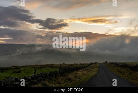 A country road leads towards a bright but cloudy late afternoon sky in Autumn in the North Pennines Stock Photo