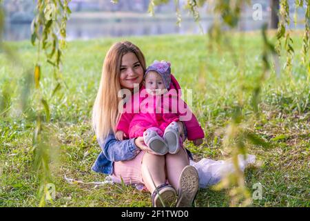 Happy young mother with her baby is sitting on the grass in the park in the autumn evening. Stock Photo