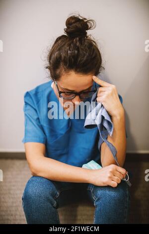Portrait of middle age woman surgeon sitting on the floor leaned on the wall after an operation and praying by looking down. Stock Photo