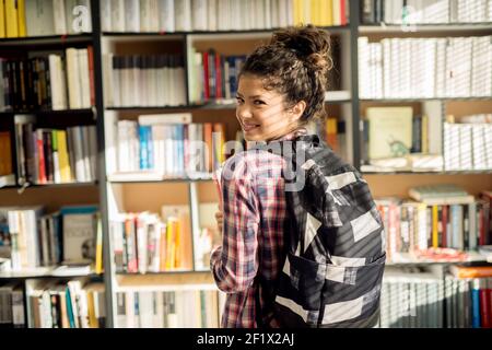 Rear view of a young gorgeous student girl with a black backpack while looking at the camera in front of the library bookshelf. Stock Photo
