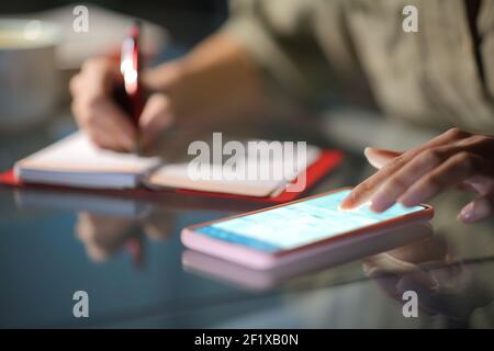 Close up of a woman hands checking smart phone calendar and writing on agenda in the night at home Stock Photo