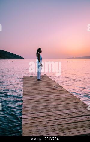 Crete Greece, young romantic couple in love is sitting and hugging on wooden pier at the beach in sunrise time with golden sky. Stock Photo