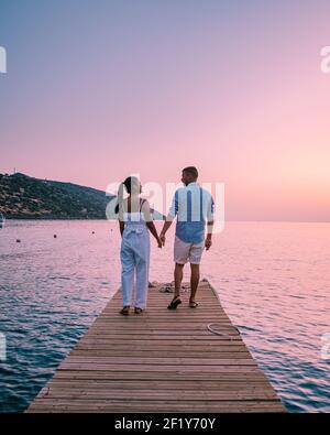 Crete Greece, young romantic couple in love is sitting and hugging on wooden pier at the beach in sunrise time with golden sky. Stock Photo