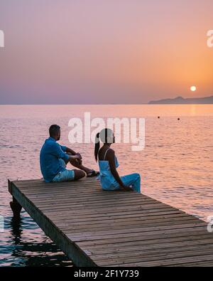 Crete Greece, young romantic couple in love is sitting and hugging on wooden pier at the beach in sunrise time with golden sky. Stock Photo