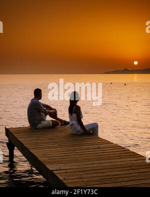 Crete Greece, young romantic couple in love is sitting and hugging on wooden pier at the beach in sunrise time with golden sky. Stock Photo