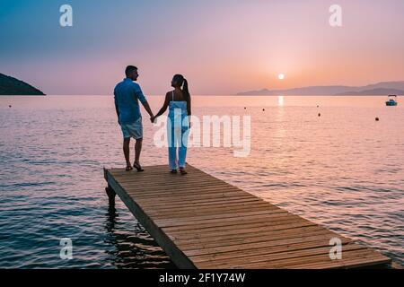 Crete Greece, young romantic couple in love is sitting and hugging on wooden pier at the beach in sunrise time with golden sky. Stock Photo