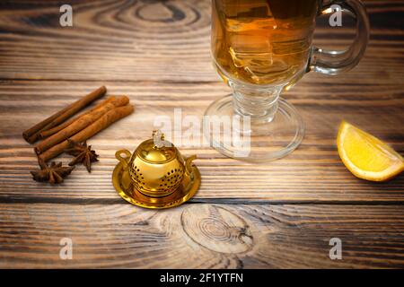 Vintage tea strainer and glass of tea on a wooden table with lemon and cinnamon. Teapot-shaped golden tea strainer. Stock Photo