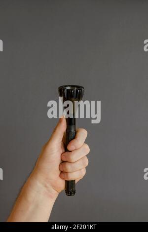 A hand holds a black modern LED flashlight on a gray background. Male hand and a metal flashlight. Side view. Selective focus. Stock Photo