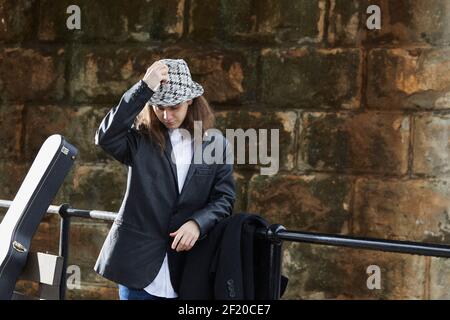 Young adult standing by a railing in front of a stone wall, adjusting a patterned hat, with a guitar case visible. Stock Photo