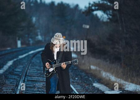 Young man in hat and coat standing on rails and playing electric guitar in evening in countryside Stock Photo