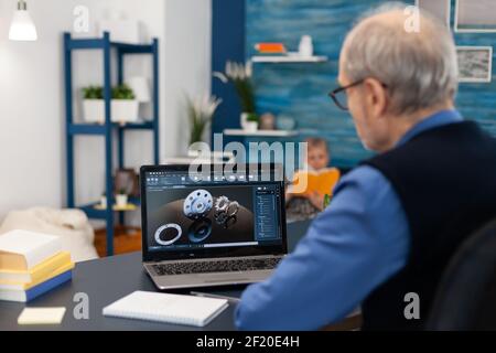 Senior engineer working on deadline sitting at desk. Experienced engineer working from home office on mechanical project using laptop computer technology and wife is reading a book on sofa. Stock Photo