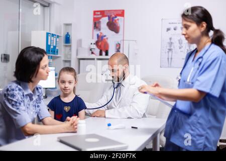Doctor pediatry checking evolution of flu using stethoscope and nurse writing disease symptoms. Healthcare practitioner in medicine providing professional treatment services in hospital clinic. Stock Photo