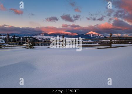 Alpine village outskirts in last evening sunset sun light. Winter snowy hills and fir trees. Stock Photo