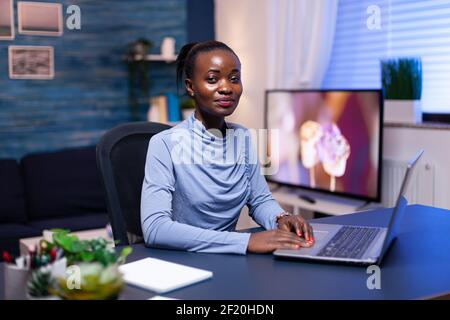 Successful african woman using laptop computer in living room late at night looking at camrea. Black entrepreneur sitting in personal workplace writing on keyboard. Stock Photo