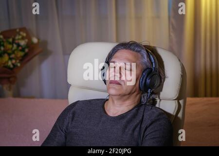 50-year-old man listens to music on headphones at home, sitting in a chair. Relaxing delight. Stock Photo