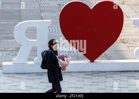 Non Exclusive: UZHHOROD, UKRAINE - MARCH 8, 2021 - A young man carries a bouquet as he walks past a huge heart-shaped installation on International Wo Stock Photo