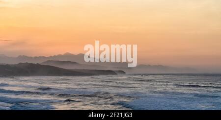 Ocean coast at sunset with waves breaking and mountains in silhouette behind Stock Photo