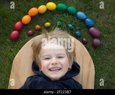 Happy toddler boy lies near chicken and quail eggs brightly painted in rainbow colors, sunny spring holiday. Family traditions. Joyful light day of Ea Stock Photo