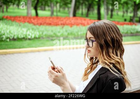 Outdoor portrait of young businesswoman with cell phone on nature background. Smartphone in hand business woman in formal suit. Stock Photo