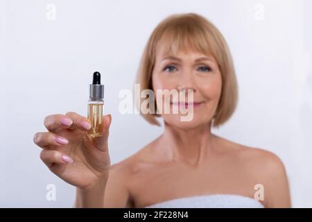 Beautiful mature woman showing bottle with rejuvenating serum on light studio background, selective focus Stock Photo