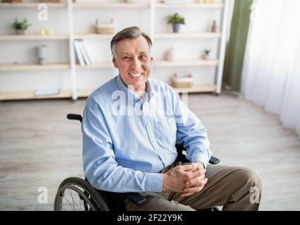 Portrait of joyful impaired senior man in wheelchair posing and smiling at home Stock Photo