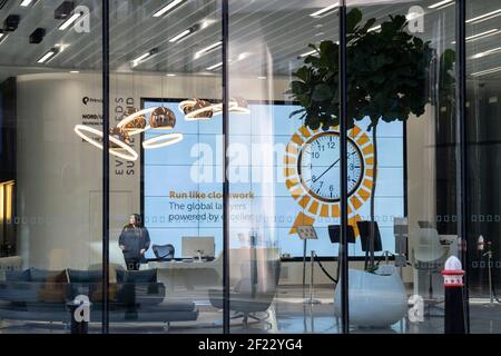 While office workers remain at home in accordance to government Covid guidelines and individual company policies, a solitary receptionist works alone beneath a clock on a screen in the City of London, the capital's financial district, during the third lockdown of the Coronavirus pandemic, on 9th March 2021, in London, England. Stock Photo