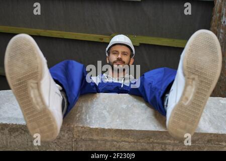 Happy handsome young male builder in blue overalls and hard hat looking at camera, sitting on the concrete floor while working on house construction. Building house concept. Low angle view Stock Photo