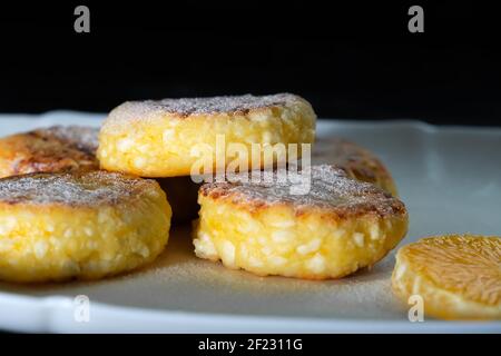 Sweet cottage cheese pancakes with orange slices on a white plate close-up against a black wall with copy space. Stock Photo