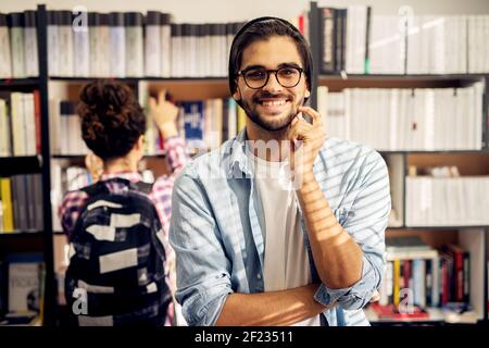 Portrait of a young cheerful male student in library scratching his beard and looking at the camera while a young joyful female student is taking a bo Stock Photo