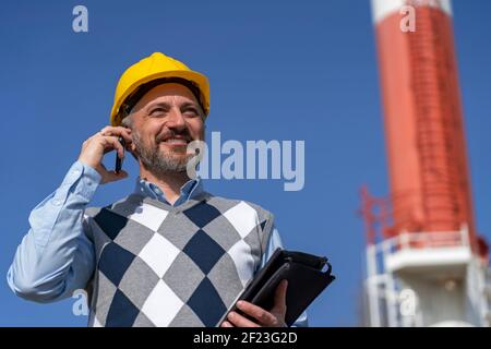 Smiling Business Person in Yellow Hardhat Talking on Mobile Phone and Standing Against Power Plant Chimney and Blue Sky Background. Stock Photo
