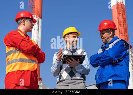 Two Workers in Personal Protective Equipment Meeting with Business Person. District Heating Power Plant. Digital Technology and Teamwork Concept. Stock Photo