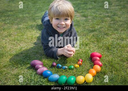 Happy toddler boy lies on the grass near chicken and quail eggs, brightly colored in rainbow colors, sunny spring day. Family traditions, joyful child Stock Photo