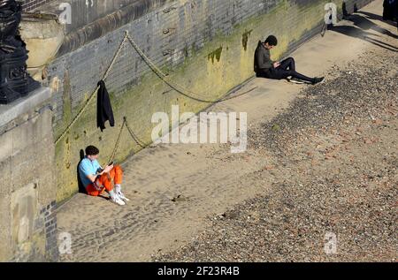 London, England, UK. Two young men on their mobile -phones sitting on the beach on the north bank of the Thames Stock Photo