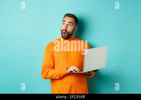 Handsome man doing online shopping, looking up thoughtful while using laptop, standing over light blue background Stock Photo