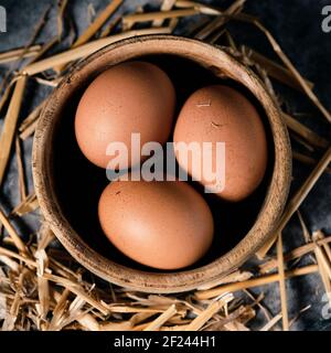 high angle view of some brown chicken eggs in a boxwood bowl and some straw stalks on a gray rustic stone surface Stock Photo