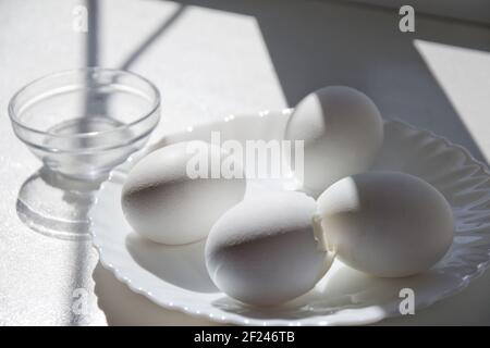 four boiled eggs in elegant little white porcelain plate. One is slice open. on clean windowsill in the sunlight Stock Photo