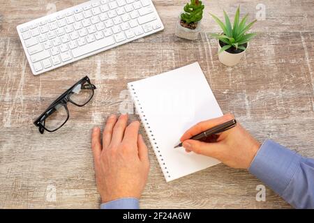 Hand of man writing in white spiral notepad on wooden office desktop with keyboard and glasses. Business report,Top view Stock Photo