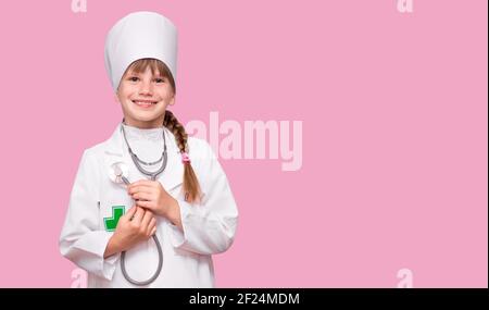 Little, cute girl future doctor dressed white medical apron with stethoscope isolated on pink background, copy space Stock Photo