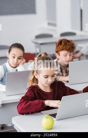 concentrated schoolgirl using laptop near apple and classmates on blurred background Stock Photo