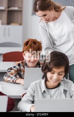 teacher standing near smiling redhead boy using laptop during lesson Stock Photo