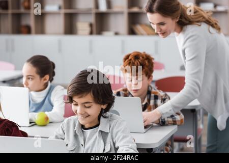 teacher helping schoolboy working on laptop during lesson, blurred background Stock Photo