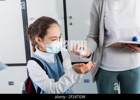 Teacher with infrared thermometer and notebook pouring sanitizer on hands of pupil in medical mask Stock Photo