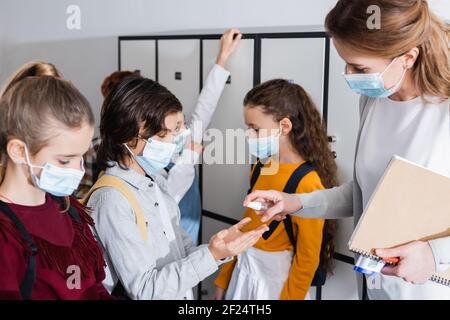 Teacher with notebook and infrared thermometer holding sanitizer near hands of pupils Stock Photo