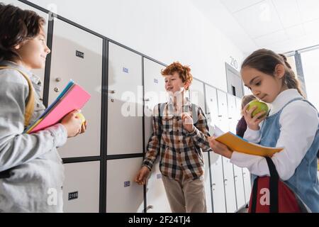 Cheerful boy with backpack standing near lockers and friends with apples and notebooks on blurred foreground in school Stock Photo