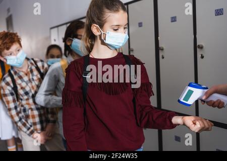 Schoolgirl in protective mask standing near teacher using non contact thermometer and classmates on blurred background Stock Photo