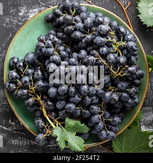 Black grapes in plate with vine plant on dark table top view. Sweet juicy grapes food on black concrete background flat lay square. Stock Photo
