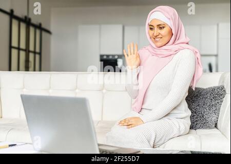 Young pretty Muslim Arabic lady in hijab sitting on the couch at home and waving at laptop, video calling, using computer for online consultation or remote communication, smiling and greeting a friend Stock Photo