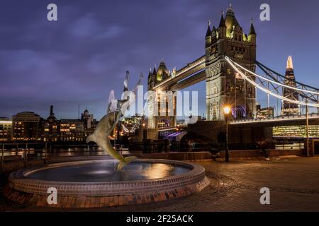 The beautiful statue of a 'Girl with a Dolphin', by David Wynne, illuminated by streetlights, against the backdrop of London's iconic Tower Bridge jus Stock Photo