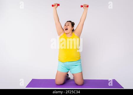 Happy plus size woman in bright home clothes doing exercises with blue dumbbells on her knees on a yoga mat. Isolated on gray background with side Stock Photo